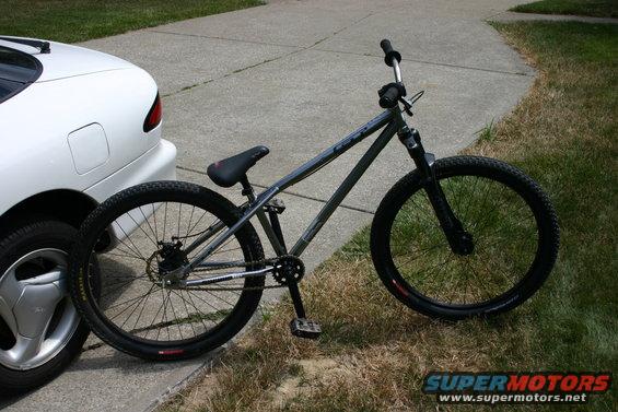 Gray dirt jump bike next to white car. Supermotors.net logo visible