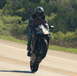 2006 Honda CBR-RR - A rider on a black motorcycle performs a wheelie on a road, with a wooded area visible behind them.