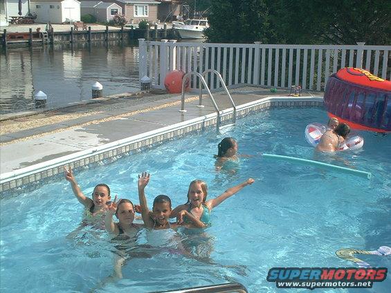 Children play in a backyard swimming pool with a canal and houses in the background. Water toys are visible