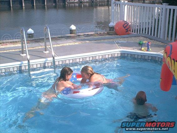 Kids swim in a backyard pool by a canal. One kid is on an inflatable ring