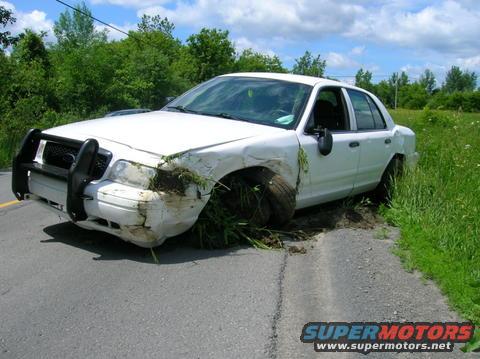 White sedan with a push bar and significant damage to the front right fender and wheel, partially off the road in grass