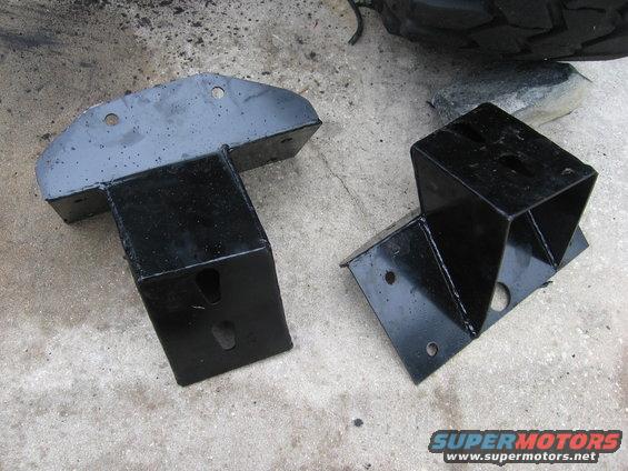 Two black metal brackets on concrete, one with a wide base and a T-shaped top, the other a cube with a base