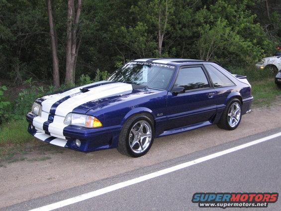 Blue 1987 Ford Mustang with white racing stripes and chrome wheels parked on a roadside