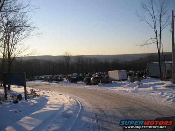 snowy parking lot with many cars and trucks, some trailers, and buildings