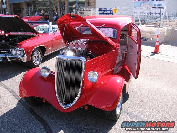 Red hot rod with hood and door open, showing exposed engine and interior. Another red car is parked to the left