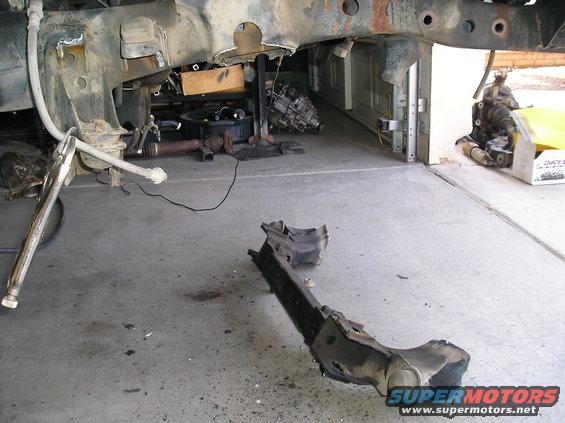 Vehicle frame and suspension components on a concrete floor, some parts appear rusted and disassembled