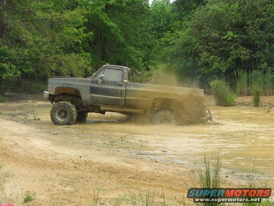 Side view of a lifted, dark-colored pickup truck splashing mud while driving through a muddy area