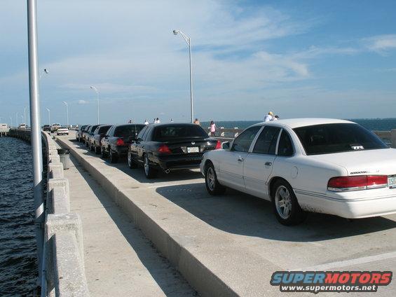 Line of black and one white 1997 Ford Crown Victoria sedans parked on a bridge, water on the left, sky above
