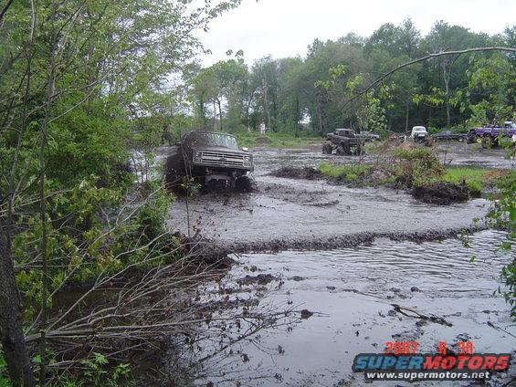 Muddy trucks driving through a swampy area. Other vehicles are visible in the background
