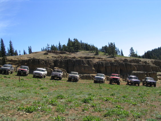Several off-road SUVs parked in a line on a grassy hill with a rocky cliff face behind them