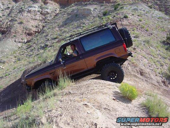 1986 Ford Bronco II driving up a steep dirt incline with a roof rack and spare tire on the back