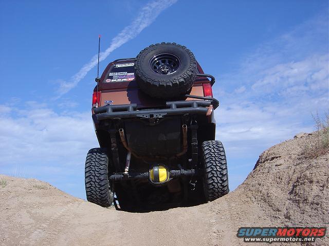 Rear view of a lifted, brown SUV with a spare tire on the back, driving on a dirt trail