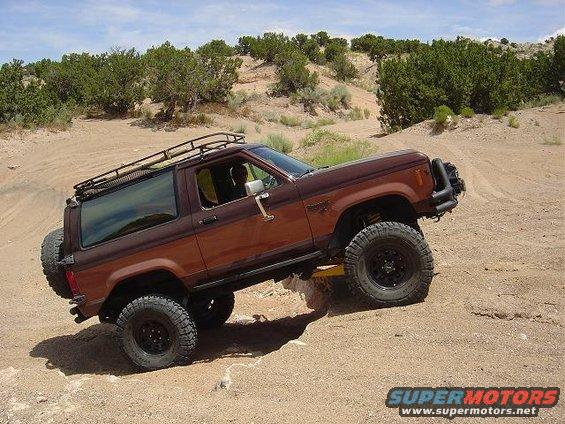 Brown 1986 Ford Bronco II climbing a dirt hill with a roof rack and spare tire on the back