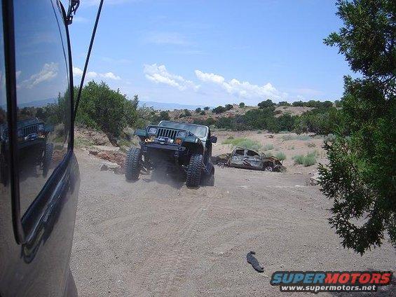 Green Jeep Wrangler with large tires driving on a dirt road, kicking up dust. A burnt-out car is visible in the background