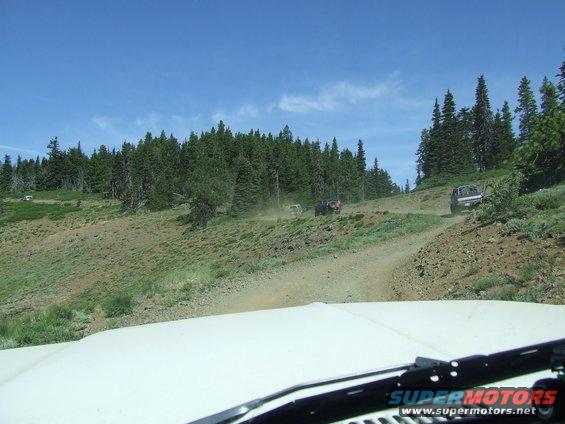 White vehicle hood in foreground, dirt road with multiple SUVs driving on a forested hillside under a blue sky