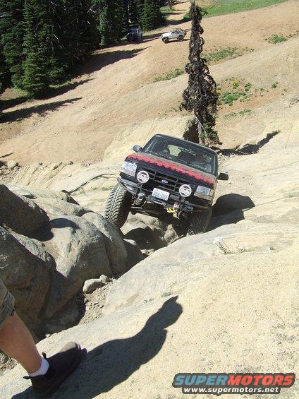 Red SUV with off-road tires navigating a rocky incline. Two other SUVs are visible further up the trail