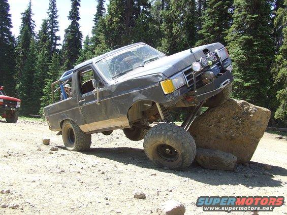 Dark gray SUV with large tires climbing over a rock, front right tire off the ground. Another red SUV is visible to the left