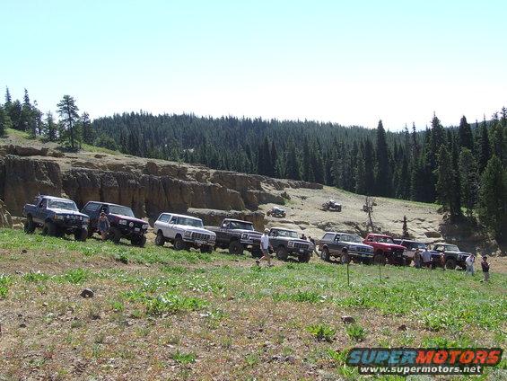 Several SUVs and trucks parked on a hillside with trees in the background. One white SUV appears to be a 1978 Ford Bronco