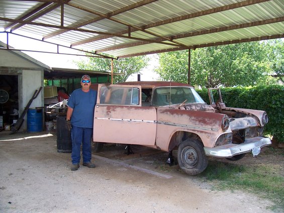 Man stands next to a rusty 1955 Ford sedan with doors open, under a metal carport