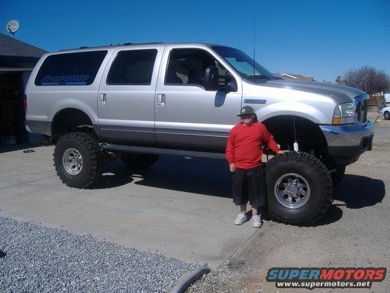 Silver lifted Ford Excursion with large tires, person in red shirt next to front tire