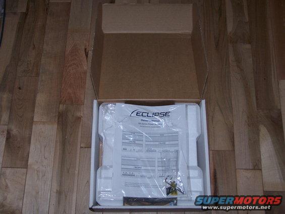 Open box containing an Eclipse car stereo owner's manual and accessories on a wooden floor