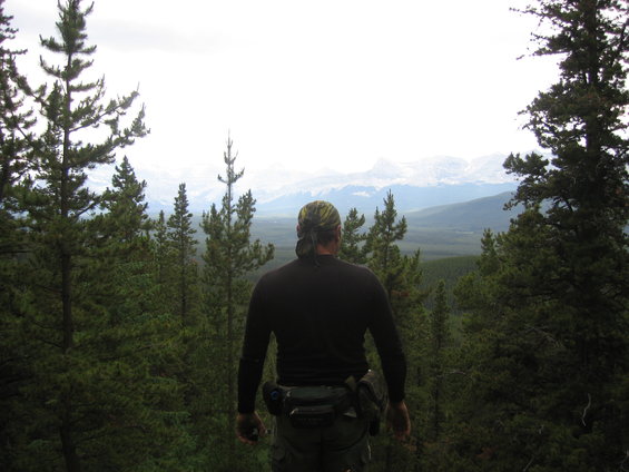 Man with bandana and fanny pack looks out over pine forest towards snow-capped mountains