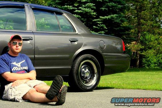 Man in blue shirt and shorts sits on the ground next to a 2000 Ford Crown Victoria