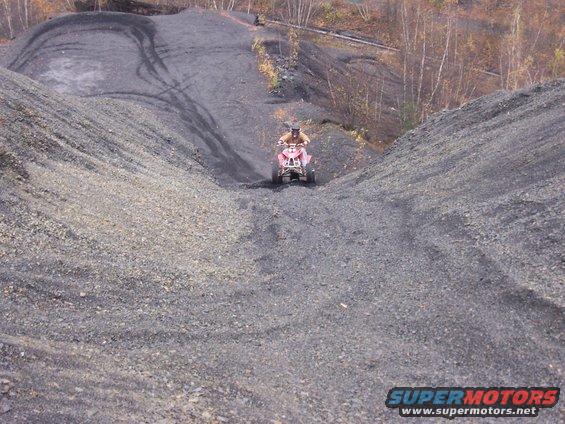 Person riding a pink ATV down a steep, loose gravel hill. Trees and industrial structures visible in the background