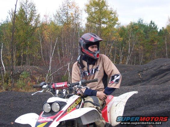 Guy in helmet and riding gear sits on a red and white quad bike. Trees and dirt mounds in background