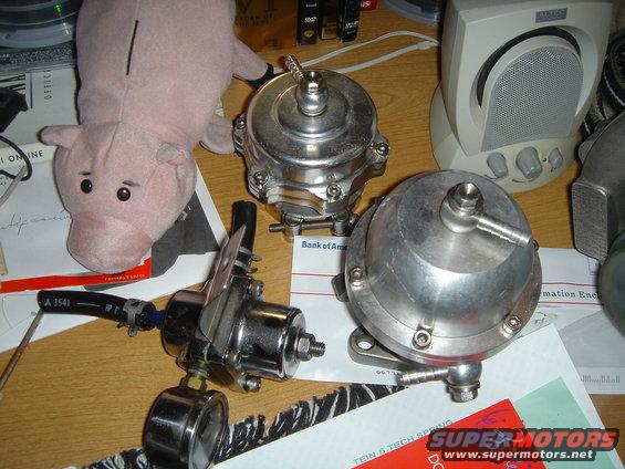 three shiny metal automotive parts, one with a gauge, and a pink stuffed pig on a desk with papers