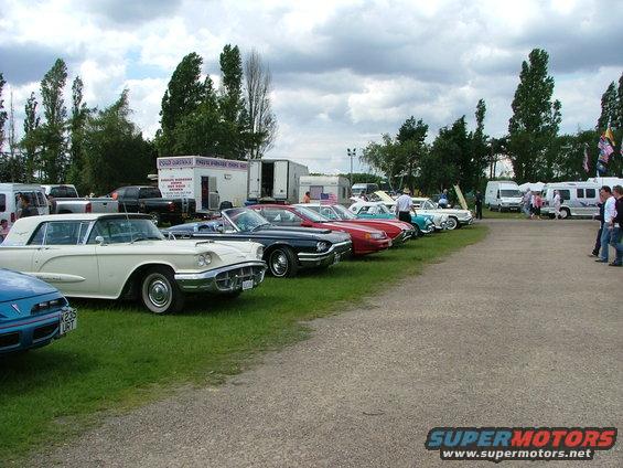 Row of vintage cars including a white convertible and a blue sedan, parked on grass with trailers and people in the background