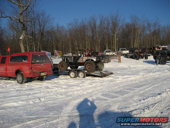 Off-road buggy on a trailer being towed by a red pickup truck in a snowy field with trees in the background