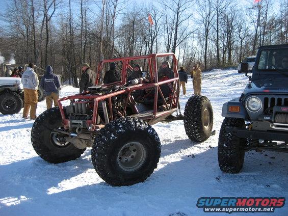 Custom red tube-frame buggy with large tires parked in snow next to a dark blue Jeep