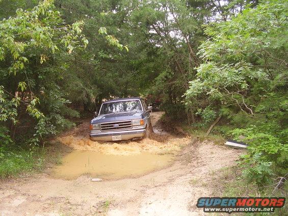 Blue Ford F-Series truck driving through a muddy puddle on a dirt road