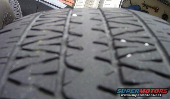 Tire tread with small white debris embedded in the grooves. A Supermotors.net logo is visible in the lower right corner