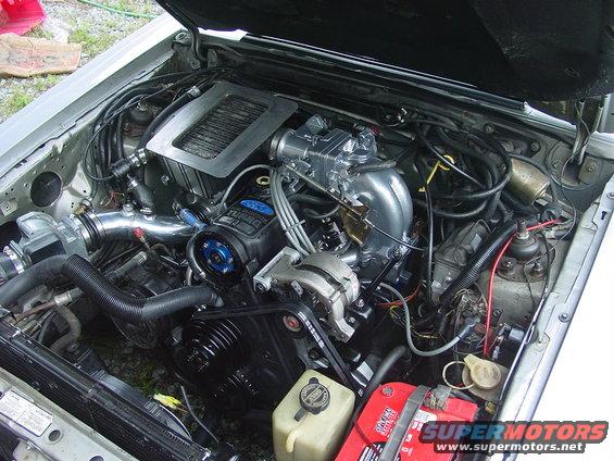 1986 Ford Mustang engine bay with silver intake, black turbocharger, and red battery