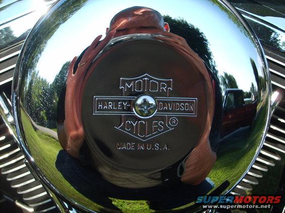 Chrome Harley-Davidson engine cover with "MOTOR HARLEY-DAVIDSON CYCLES MADE IN U.S.A." text. Reflection shows a person and a red truck