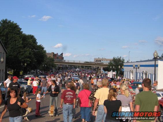 Crowded street with people and parked cars under a blue sky with clouds