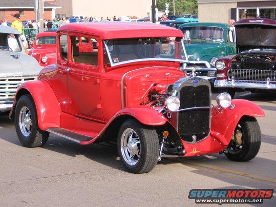 Red 1930s coupe hot rod with chrome grille and exposed engine, parked outdoors