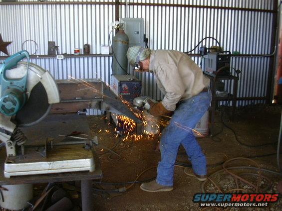 Man in workshop grinding metal with sparks flying. Chop saw and welding equipment visible