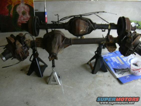 Two rusty vehicle axles are propped up on jack stands in a garage. A painting is on the wall behind them