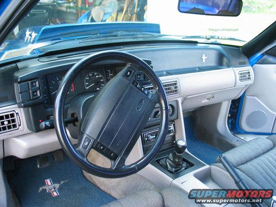 Interior of a 1990 Ford Mustang with a blue dashboard and grey seats. Steering wheel, gear shift, and radio are visible