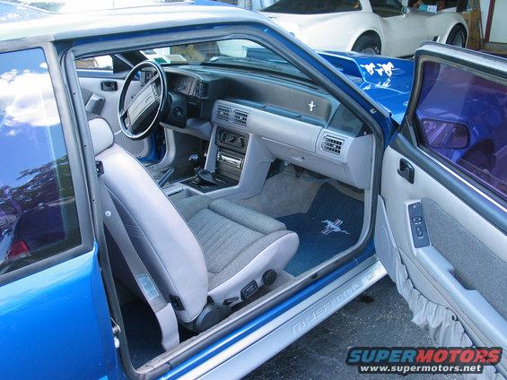 Interior of a 1990 Ford Mustang, showing dashboard, seats, and door panel. Another car is visible in the background