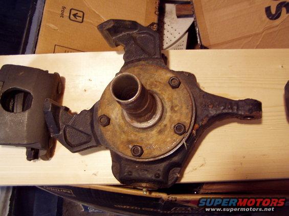 Vehicle axle assembly and brake caliper on a wooden surface. The axle has a rusty hub and is bolted to a bracket