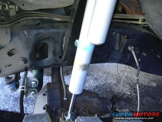 Close-up of a white Bilstein shock absorber attached to a black metal frame with visible brake lines and suspension components