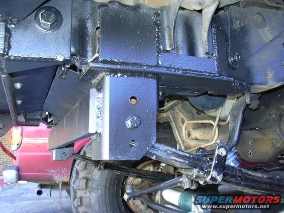 Close-up of custom metal brackets welded to a vehicle's frame, with suspension components visible below. A red vehicle is partially seen in the background