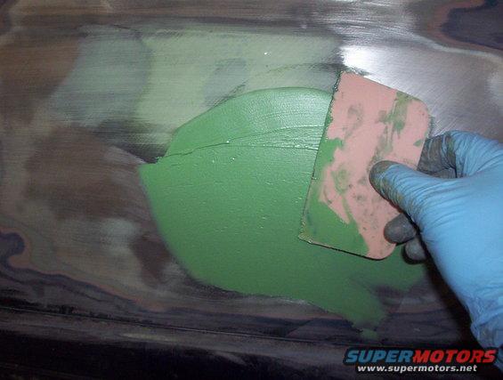Hand in blue glove applying green body filler with a pink spreader to a metal surface