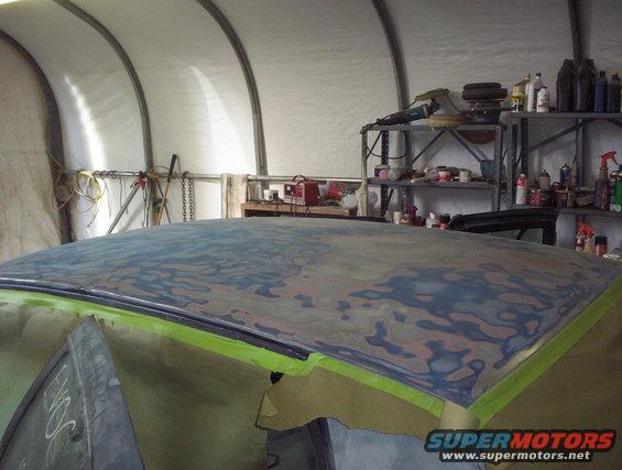Car roof covered in primer and body filler, taped off for painting. Tools and supplies on shelves in background