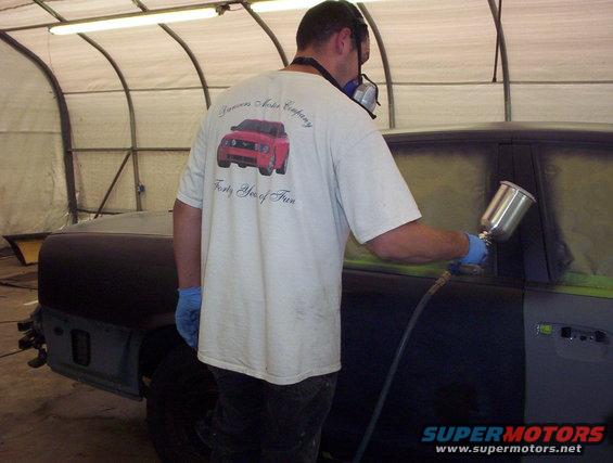 Man in respirator and gloves sprays paint onto a car door in a spray booth