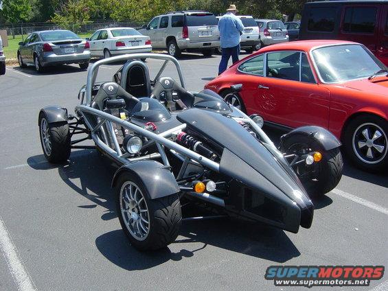 Exposed frame sports car with a dark grey body kit and two seats. Red Porsche in background